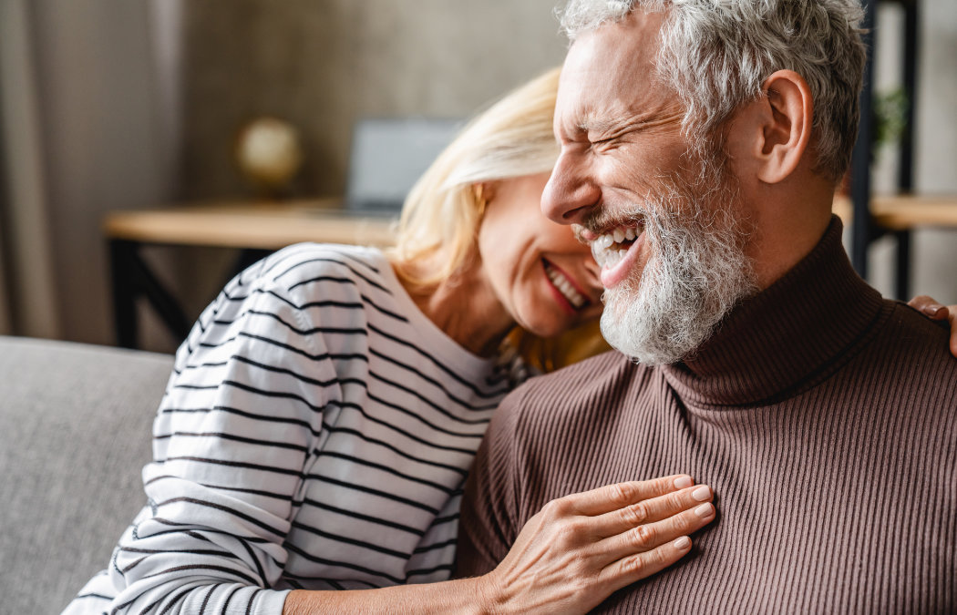 An older woman and man sit close together, smiling and laughing. The woman wears a striped shirt; the man has a gray beard and wears a brown turtleneck sweater.