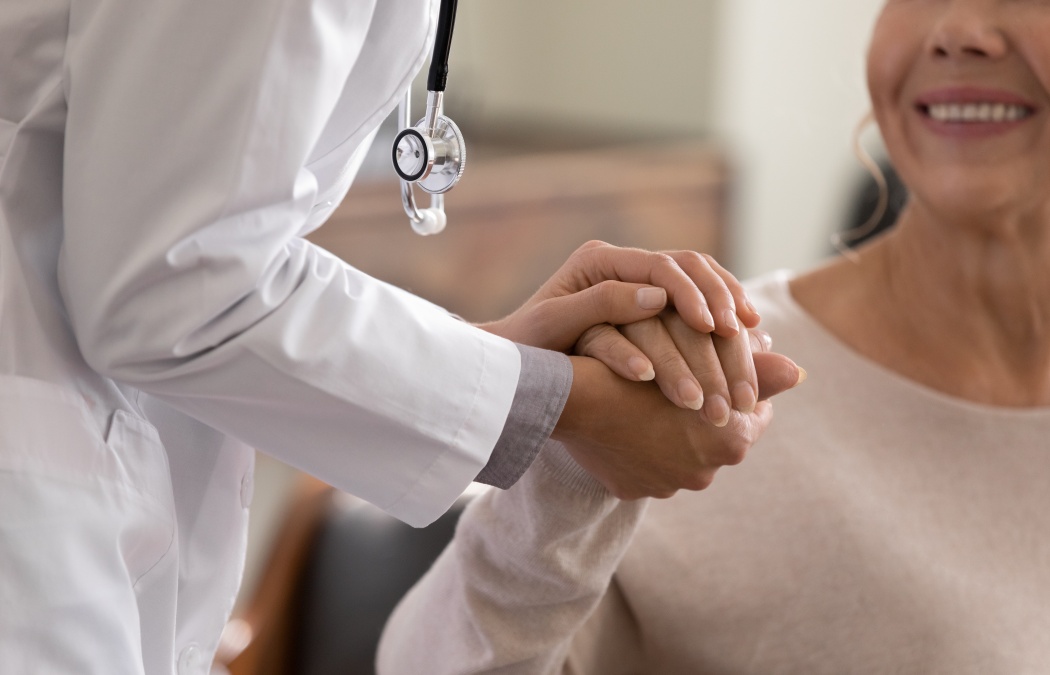 Doctor giving hope. Close up shot of young female physician leaning forward to smiling elderly lady patient holding her hand in palms. Woman caretaker in white coat supporting encouraging old person