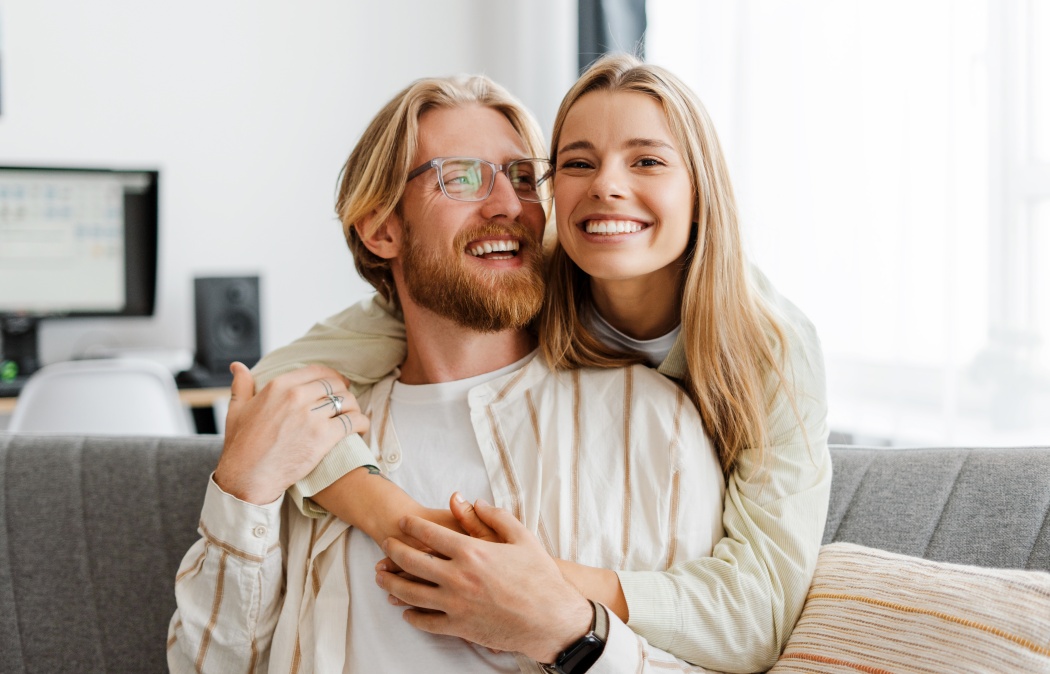 Young couple embraces on their cozy sofa at home, both smiling and radiating love and happiness in their modern living room. Relationship concept